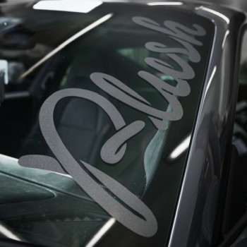 A large, cursive "Flush" decal is adhered to the windshield of a dark-colored car in a dimly lit garage, with the interior and surrounding vehicles slightly visible in the background.