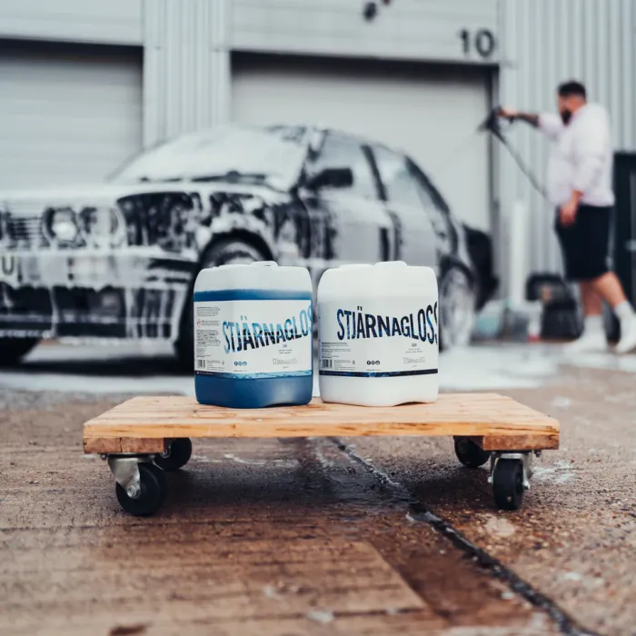 Two containers of "STJÄRNAGLOSS" cleaning product on a wheeled wooden cart; a foamy car in the background being pressure washed by a person in an industrial setting.