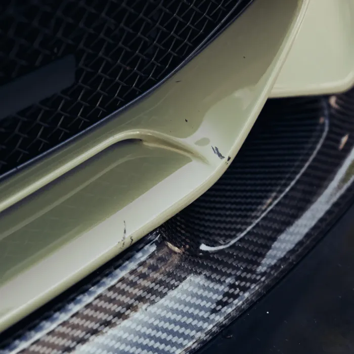 Bumper of a light-colored car with scratches sits above a carbon fiber splitter, adjacent to a dark mesh grille.
