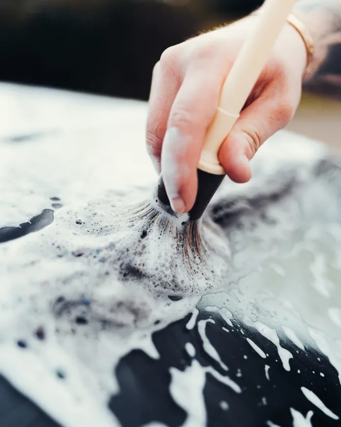 A hand using a brush creates suds on a dark, glossy surface, likely a car, in an outdoor setting.