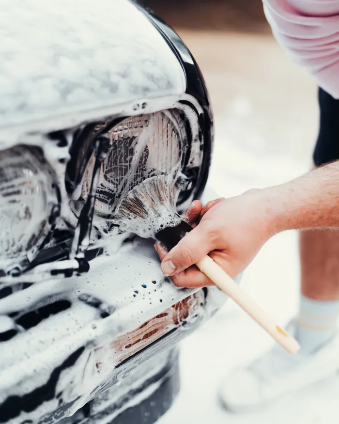 A hand uses a paintbrush to scrub a soapy car headlight, cleaning it meticulously. The surrounding area is slightly blurred, focusing on the cleaning action.