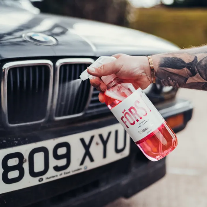 A tattooed hand sprays "FÖRS" stain remover onto the front grille of a black BMW car with license plate "809 XYU" outdoors.