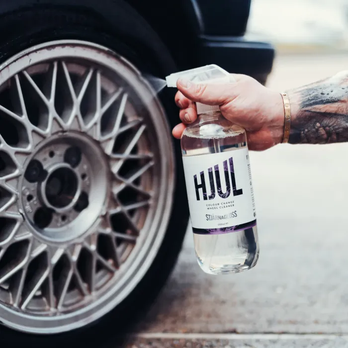 A hand is spraying a bottle of "HJUL Colour Change Wheel Cleaner" towards the rim of a car wheel on a paved surface. The car is black with detailed wheel design.