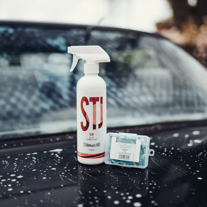 A white spray bottle labeled "STJ GLIR CLAY LUBRICANT STJÄRNAGLOSS 500ML" and a small clear plastic box rest on a soap-splattered car hood and windshield.