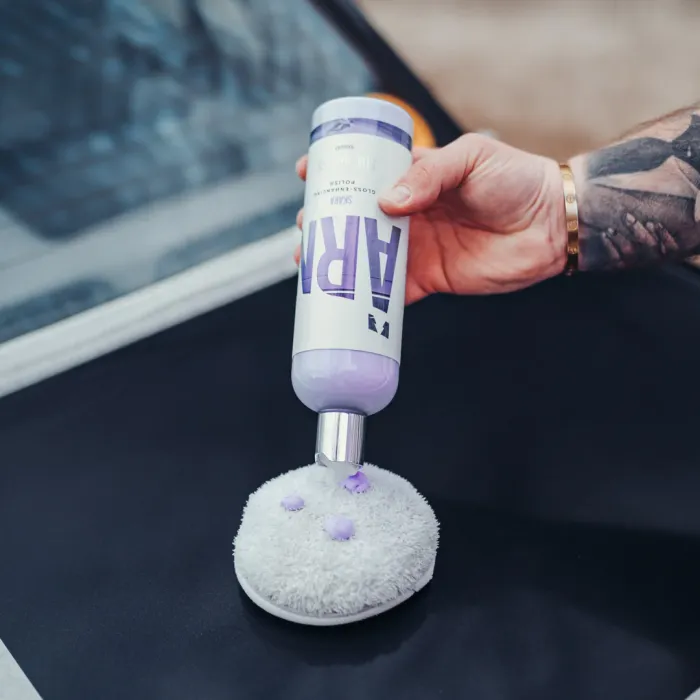 A hand holding a bottle labeled "ADBL QAR 03" dispenses purple liquid onto a white, fluffy applicator pad. A car windshield and metallic surface are visible in the background.
