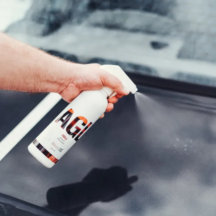A hand sprays a bottle labeled "AQUA" onto a surface in an outdoor setting, presumably cleaning a car.
