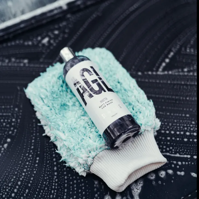 A bottle labeled "MATTA Matt Finish Car Wash" lies on a light blue, shaggy cloth on top of a soapy car surface in a car wash setting.
