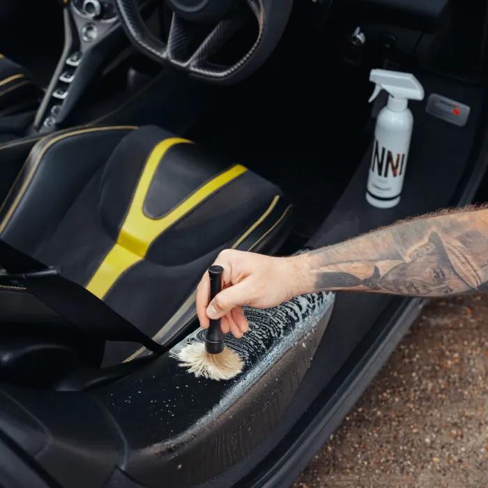 A hand uses a brush to clean a car's interior door sill, with foam visible; a spray bottle labeled "NNI" and a car seat with yellow and black upholstery are present.