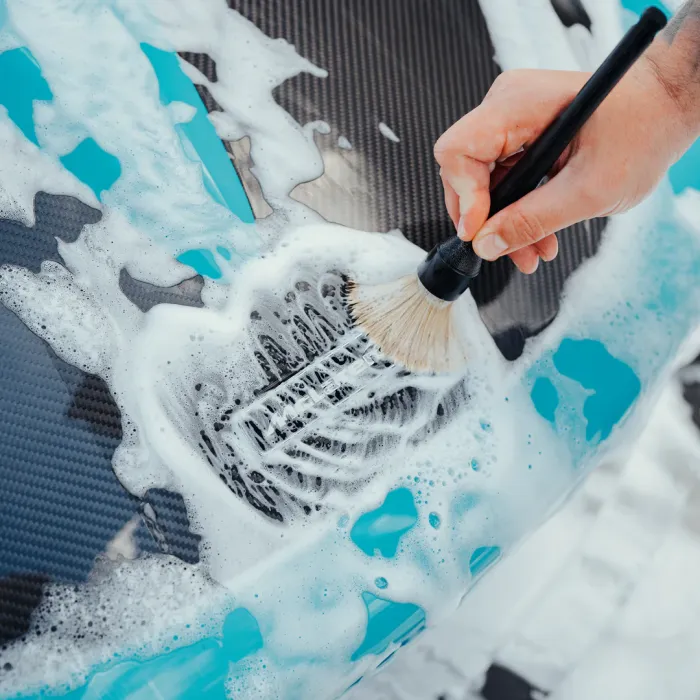 A hand holding a brush scrubs a soapy, foam-covered, black carbon fiber car part in a cleaning context.