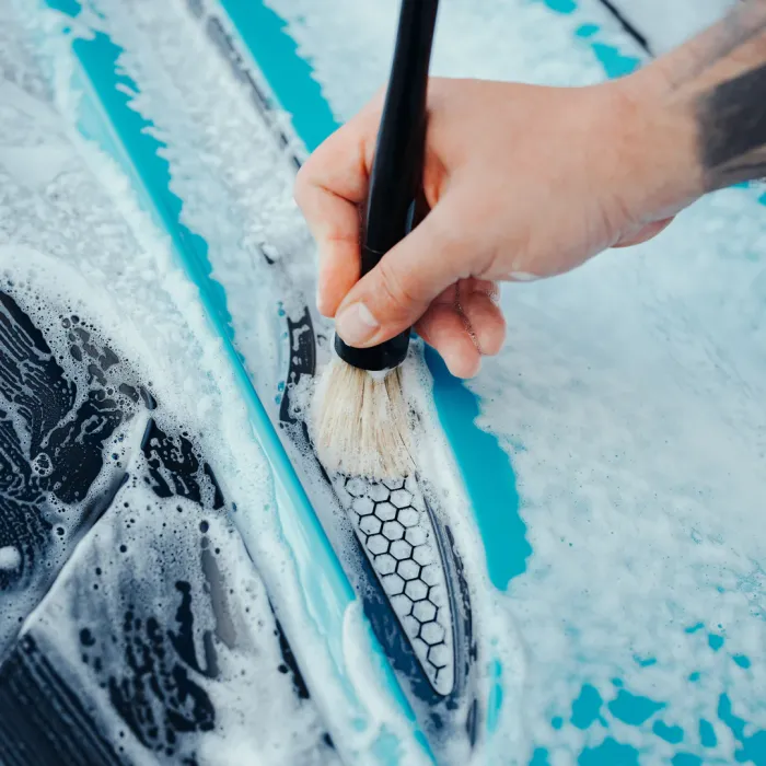 Hand holding a brush scrubbing a soapy, hexagon-patterned surface of an object (likely a car) with light blue and white foam covering much of the dark, glossy exterior.