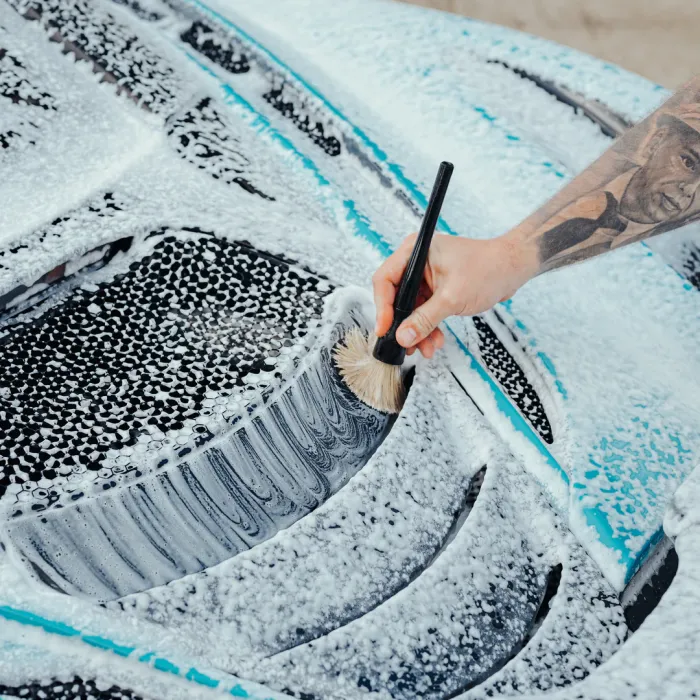 A hand with a tattoo holds a brush, cleaning a soapy car hood with a detailed black vent, amidst suds and foam in an outdoor area.