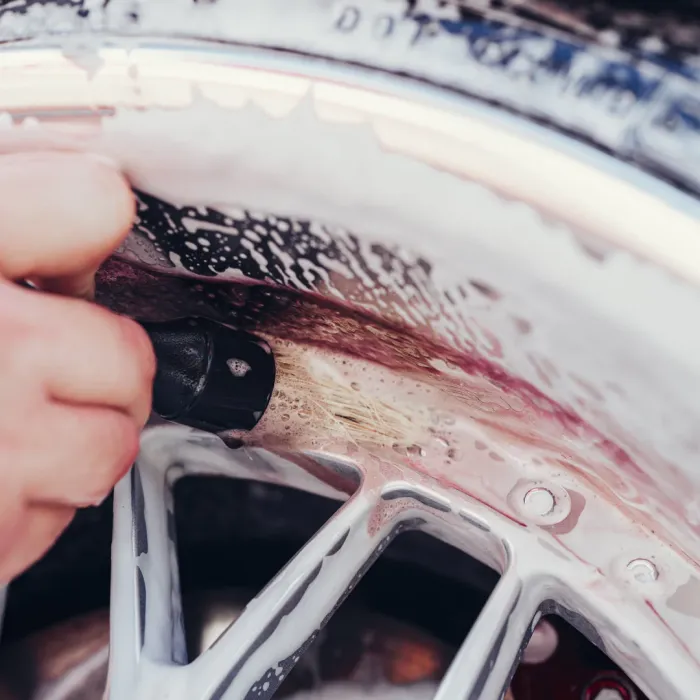 Hand cleaning a soapy car wheel rim with a brush, detailing the inner surface. The environment is a close-up view focused on the wheel and brush in use.