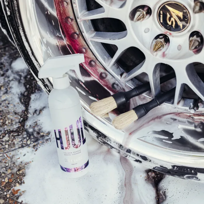 A bottle of HUL Colour Change Wheel Cleaner stands next to a vehicle's silver alloy wheel, partially cleaned with residue and suds, along with two wheel cleaning brushes lying on the wheel.