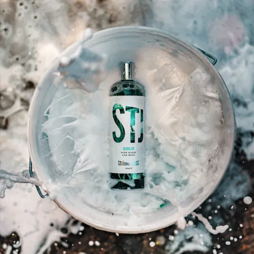 A bottle labeled "STJ" (Stjärngloss, BUBBLOR, High Gloss Car Wash, 500ml) is standing in a bucket filled with soapy water, with soap suds splashing around it.