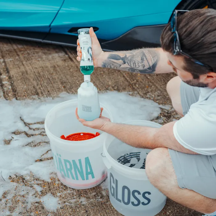 A man in gray shorts crouches beside a blue car, pouring green liquid from a bottle into a container labeled "STJÄRNA GLOSS." Two similar buckets are placed on a sudsy ground.