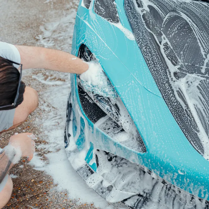 A person scrubs the soapy headlight of a turquoise car, which is covered in white foam, while kneeling on a wet, soapy ground.