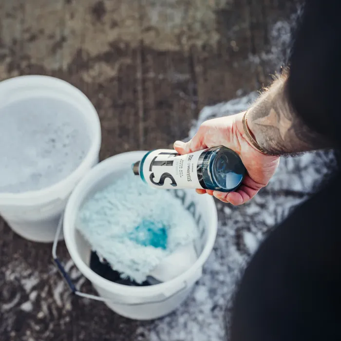 A tattooed hand holds a bottle labeled "SiO2 Ceramic Car Shampoo Quick Details," over white buckets filled with soapy water and cleaning tools, on a wet outdoor surface.