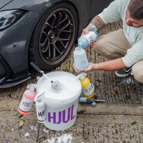 A person fills a spray bottle with car cleaning solution beside a car's wheel. Various labeled cleaning products and a foam-filled bucket on the ground include: "HJUL," "FÖRS," "GLATT."
