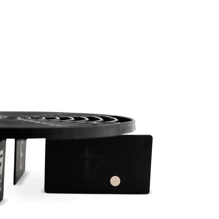 A black circular grill sits on a flat surface supported by angular metal structures, one with a triangle and dot embossed on it, set against a plain white background.