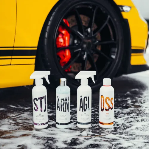 Four bottles of car cleaning products stand in foamy water before a yellow car with black wheels and red brake calipers. Labels: STJ, ÁRN, ÁGI, OSS.