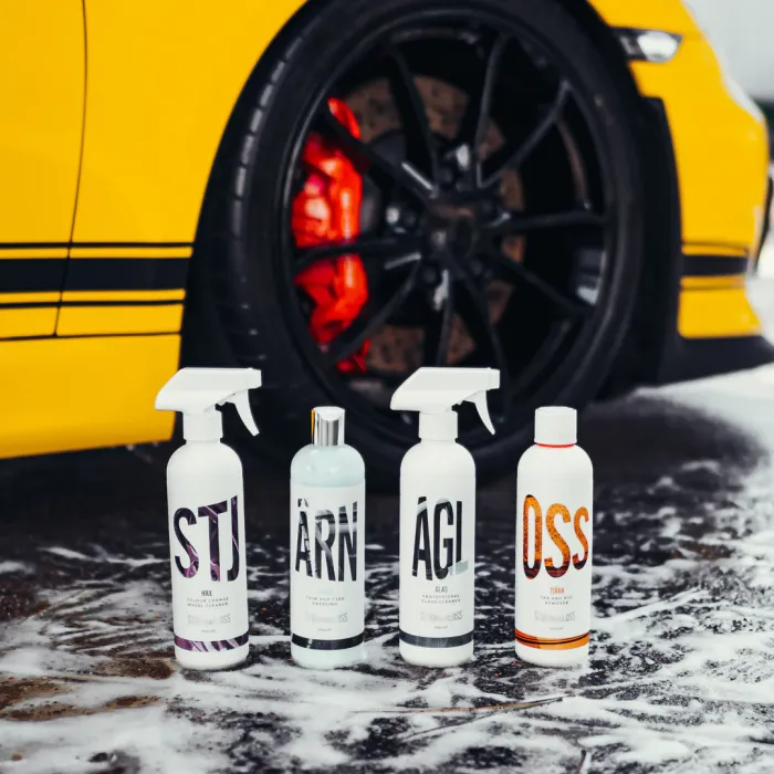 Four bottles of car cleaning products stand in foamy water before a yellow car with black wheels and red brake calipers. Labels: STJ, ÁRN, ÁGI, OSS.