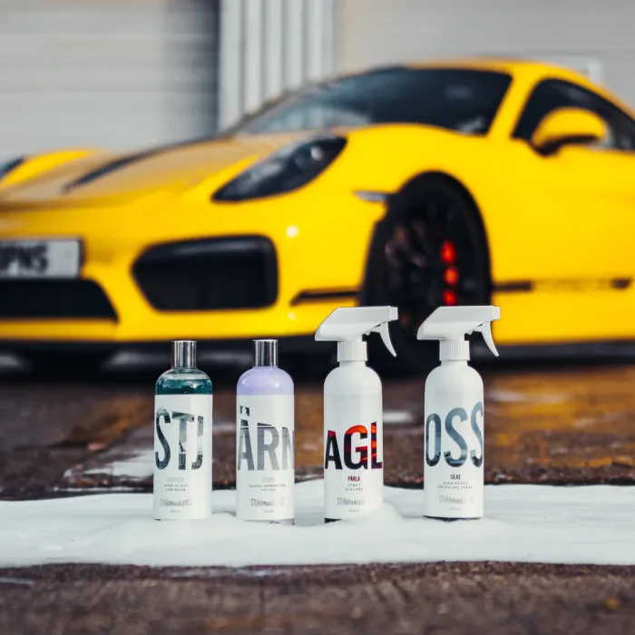 Four bottles of car cleaning products sit on a soapy ground with a yellow sports car in the background. The bottles display the following text: "STJ", "ÄRN", "AGL", and "OSS".
