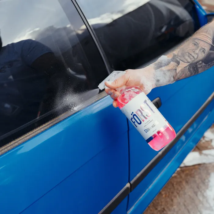 A tattooed hand sprays a pink liquid from a bottle labeled "FÖRST" onto the window of a blue car in an outdoor setting.