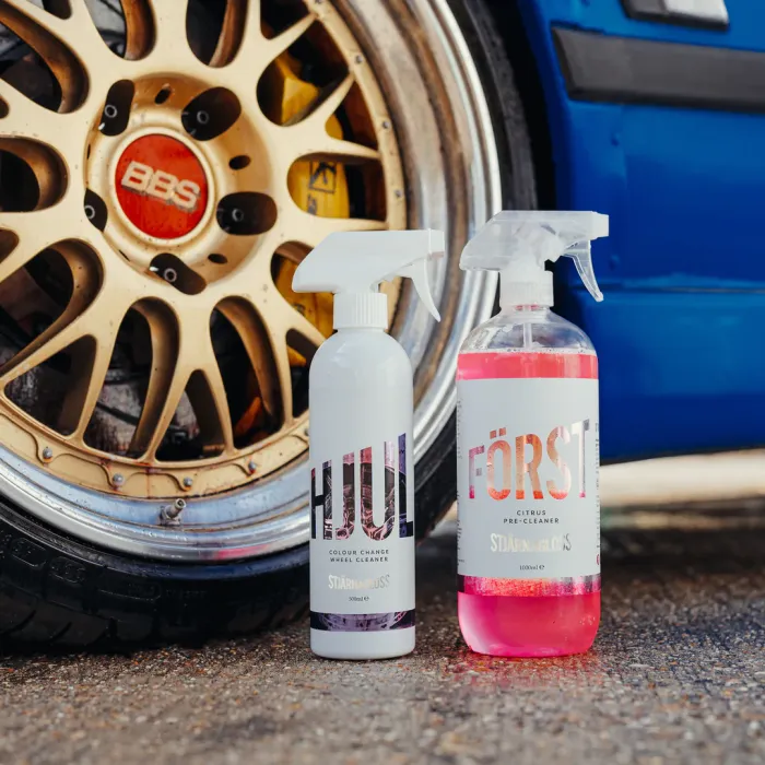 Bottles of wheel cleaner labelled "HJUL" and "FÖRST" leaning on a gold car wheel with "BBS" at its center; the blue car body is visible in the background.