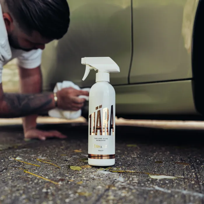 A white spray bottle labeled "TJÄRA Tar and Glue Remover" stands upright on asphalt while a person in the background, crouching, cleans a car with a cloth.