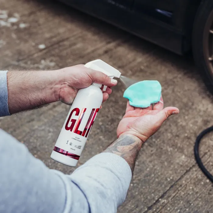 A person sprays lubricant from a bottle labeled "GLIM" onto a blue clay bar, preparing for car detailing on a concrete surface.