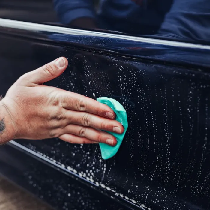 A hand with a tattoo cleans a soapy dark-colored car door using a green sponge in an outdoor setting.