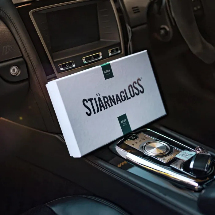 A rectangular box marked "Stjärnagloss" rests diagonally on a car's center console, featuring the gear selector, surrounded by dark leather interior.