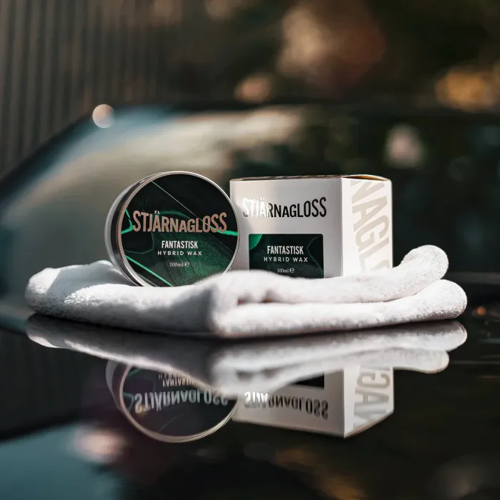 A tin of Stjärnagloss Fantastisk Hybrid Wax and its box are placed on a white towel, with a glossy car hood and blurred greenery in the background.