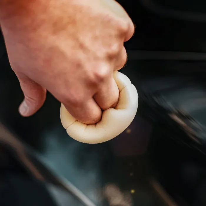 A hand grips a soft, round sponge, cleaning a dark, reflective surface, likely a car, in close-up detail.