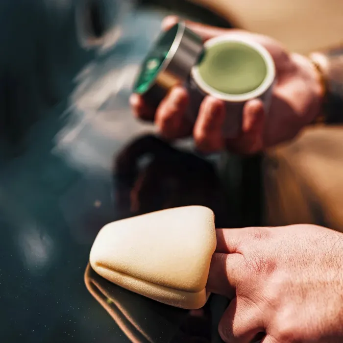A hand holds a sponge applicator against a car surface with a small container of green car wax in the background in an outdoor setting.