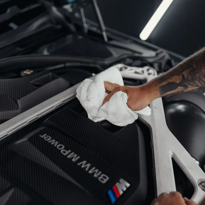 A hand with a tattoo cleans a BMW M Power engine cover with a white towel, inside a well-lit garage with structural elements visible.