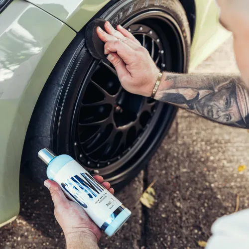 A person is applying tire dressing with a sponge to a black car tire, holding a bottle labeled "CARPRO Perl" near a parked green vehicle on an outdoor surface.