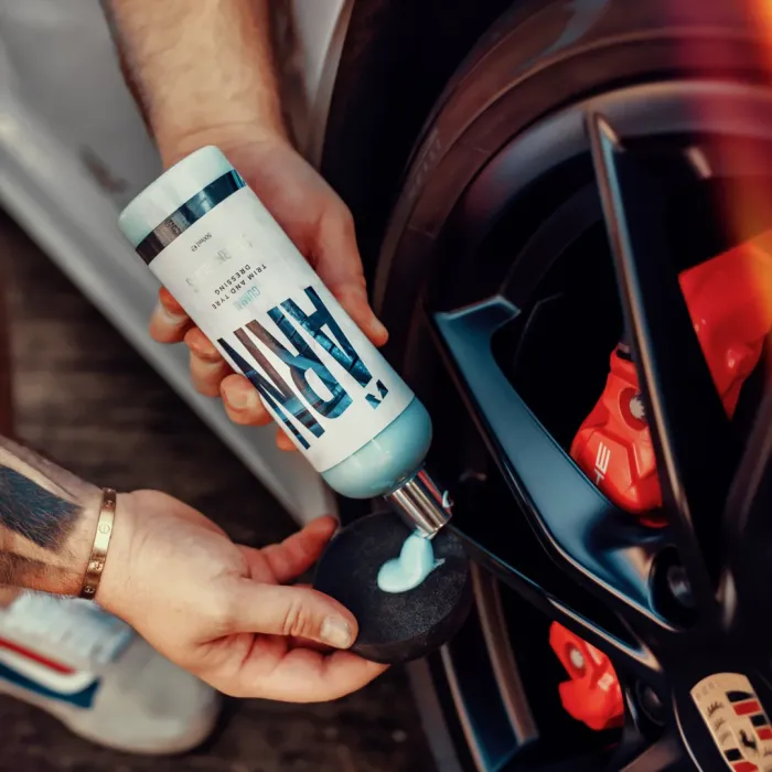 A person squeezes blue tire dressing labeled "ARMOR" onto a round black applicator sponge near a car's tire and red brake caliper.