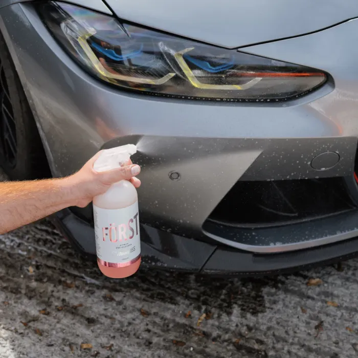 A hand sprays a solution labeled "FÖRST" onto the front bumper of a silver car in an outdoor, wet environment.