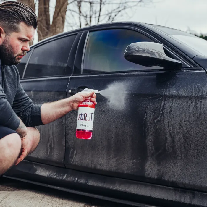 A man sprays a red cleaning solution, labeled "FÖRST" on a dirty black car door in an outdoor setting with trees in the background.
