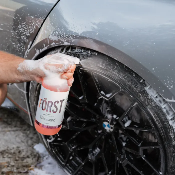 A hand sprays “FÖRST Citrus Pre-Cleanser” onto a soapy, black car wheel with a BMW logo, cleaning it in a driveway.