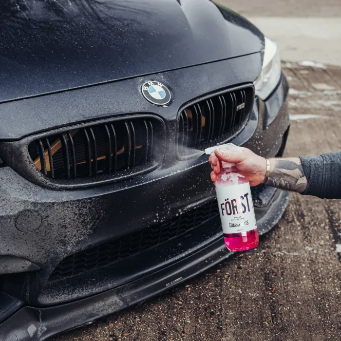 A person sprays a red cleaning solution "FÖRST CITRUS" onto the front of a dirty black BMW car, parked on a concrete surface.