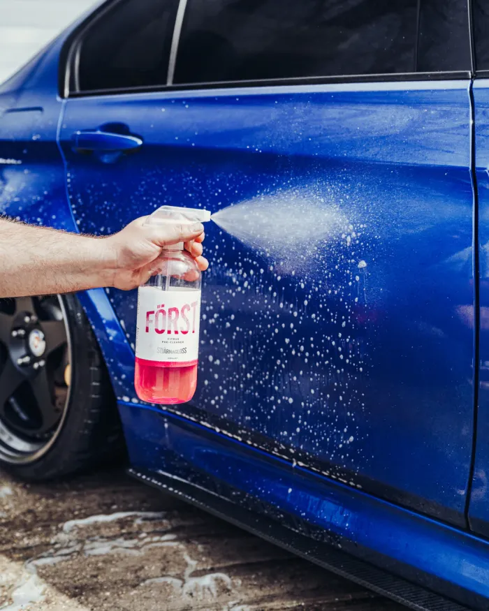 Hand sprays cleaning solution from a bottle labeled "FÖRST" onto a blue car door, which is wet and soapy. The car is parked on a wet concrete surface.