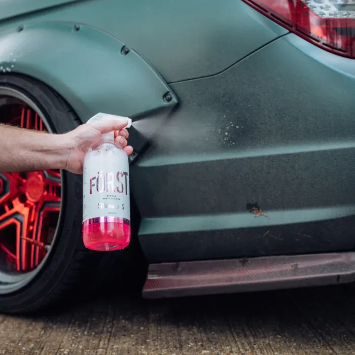 A hand sprays a pink liquid from a "FÖRST Citrus Pre-Cleaner" bottle onto the rear part of a car with red-rimmed wheels in an outdoor setting.