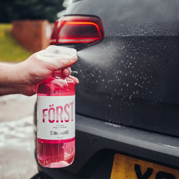 A hand sprays "FÖRST Citrus Pre-Cleaner" from a pink bottle onto a black car's rear in an outdoor setting with grass and brick nearby.