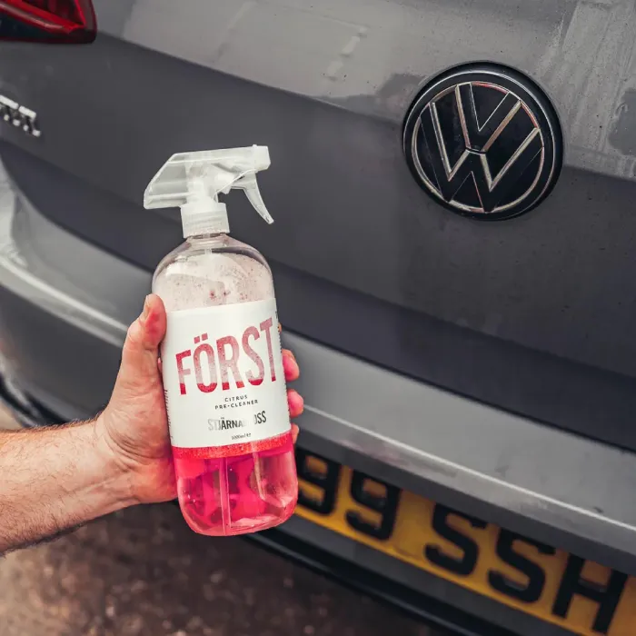 A hand holds a spray bottle labeled "FÖRST Citrus Pre-Cleaner STJÄRNAGLOSS 500ml" near the rear of a gray Volkswagen car with visible license plate, outdoors.