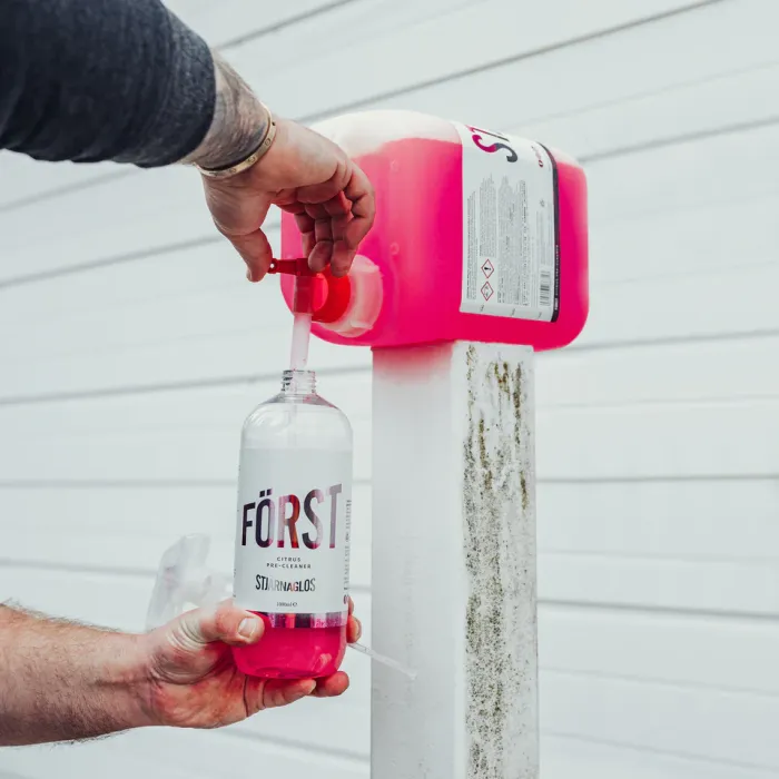 Hand fills a "FÖRST Citrus Pre-Cleaner" spray bottle from a larger pink liquid container on a white pole, with white horizontal paneling in the background.
