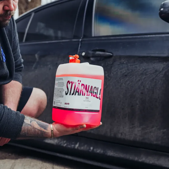 A person holds a plastic container labeled "STJÄRNAGLOSS" containing a red liquid, near a dirty, black car's door in an outdoor setting.