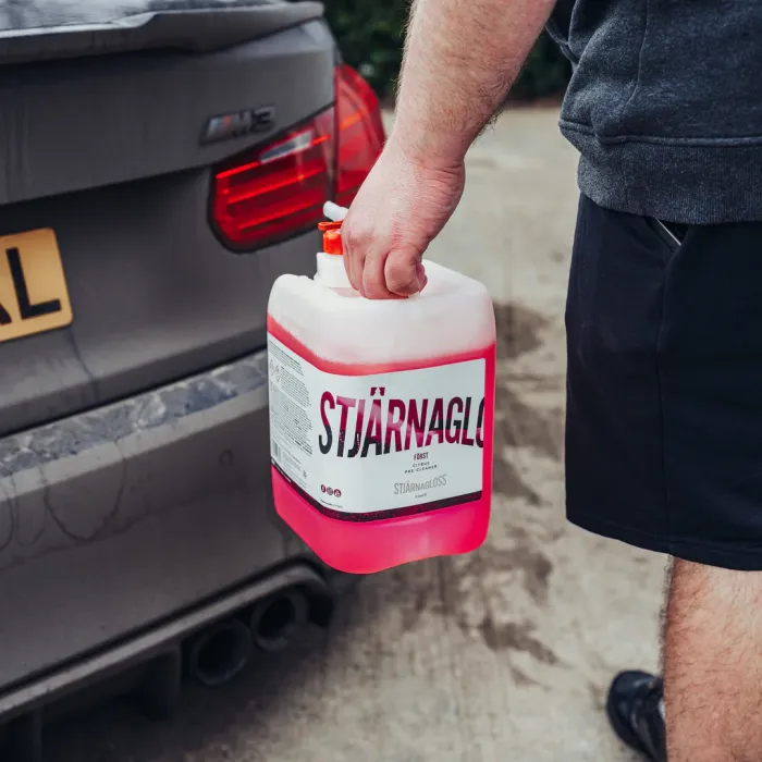 A hand holds a container labeled "STJÄRNAGLOSS" filled with pink liquid, standing next to a silver car's rear with a blurred background. Text on the container includes "FORST," "STJARNAGLOSS," and additional information.