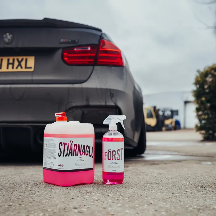 Two containers of pink cleaning solution labeled "STJÄRNAGLOSS FÖRST," positioned behind a grey BMW M3 in a parking lot, with industrial buildings and plants in the background.
